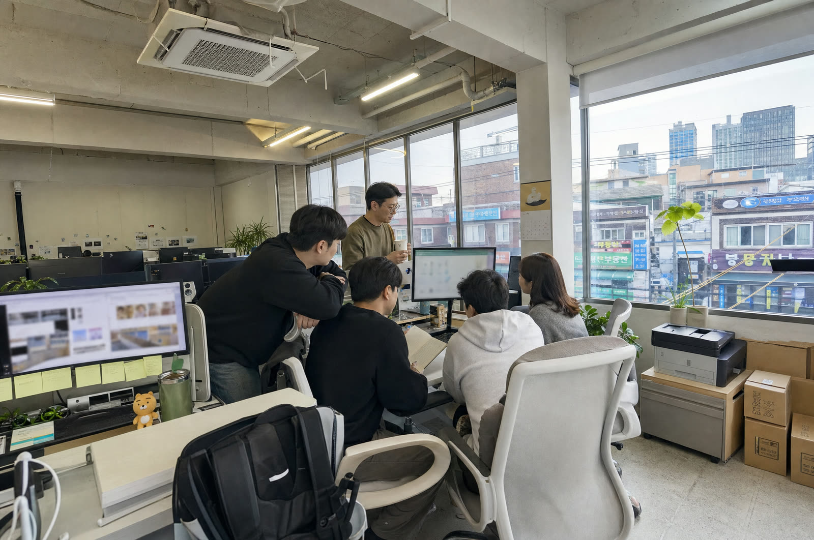 창가 자리에서 다섯 사람이 모니터 한 대를 함께 보며 이야기하는 모습. 창밖으로 거리와 간판이 보인다